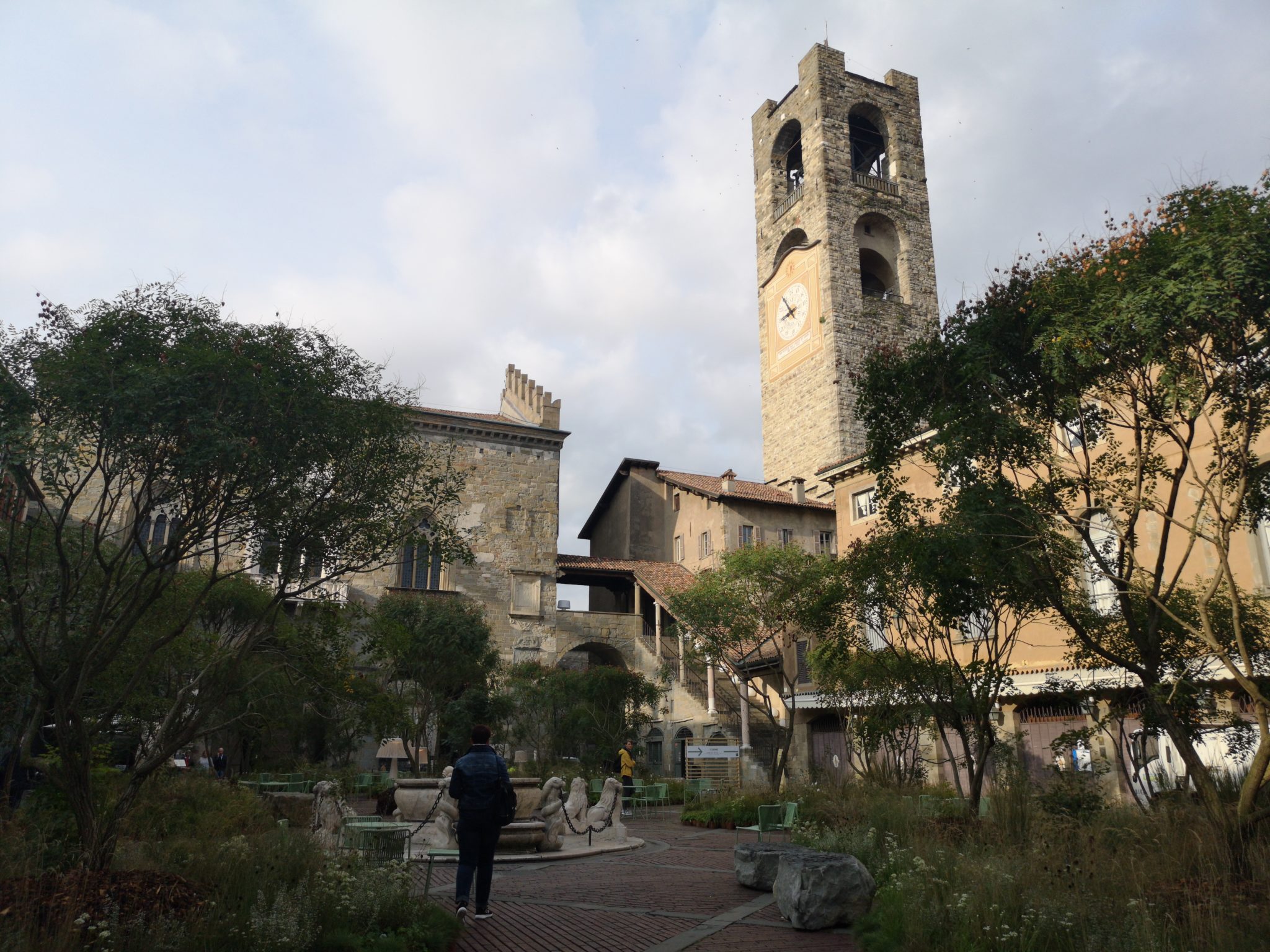 Widok na duży plac w mieście Bergamo, wysoka wieża z dzwonem i dużo zieleni, wokół kawiarnie i stoliki