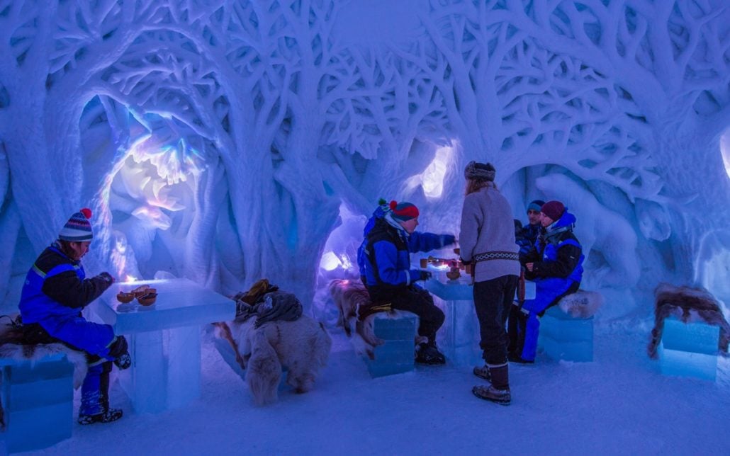 Bardzo ciepło ubrani ludzie siedzący w restauracji w hotelu lodowym pijący ze szklanek wykonanych z lodu. Ściany wrzeźbione w lodzie.